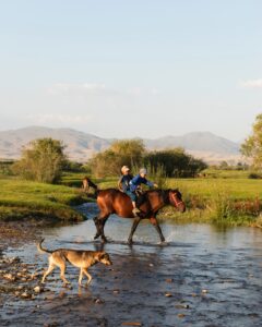 zwei Jungen reiten auf einem Pferd, begleitet von einem Hund, durch die kirgisische Landschaft.