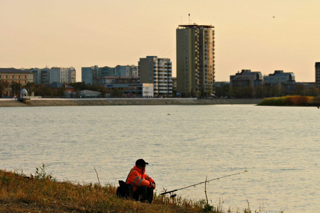 A man fishes in the Ural. He is wearing a bright orange jacket. In the background, a city, 