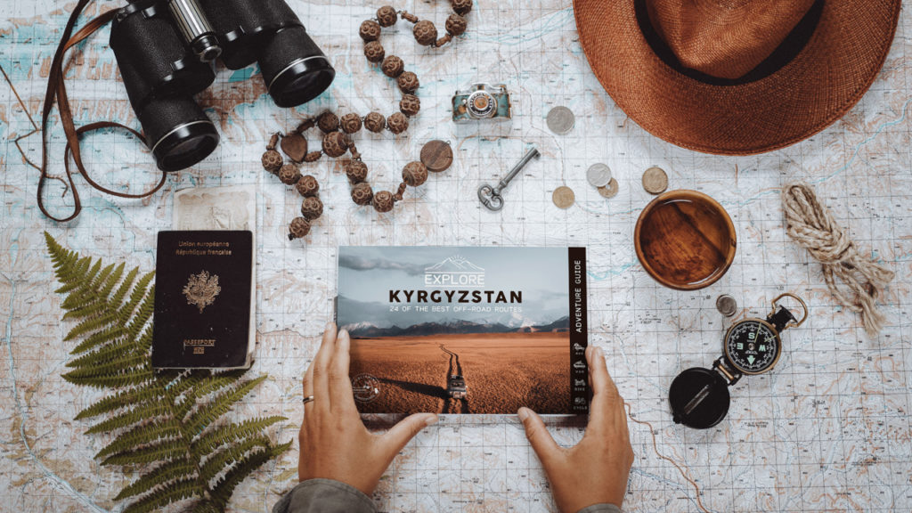 The book Explore Kyrgyzstan surrounded by a hat, a passport, binoculars, coins, a key, rope and other symbols of adventure travel