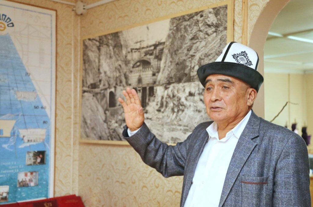 Sabirzhan Toktogulov. His hand is raised and he is wearing a kalpak. Behind him, a black and white photo of the construction of the Toktogul power station